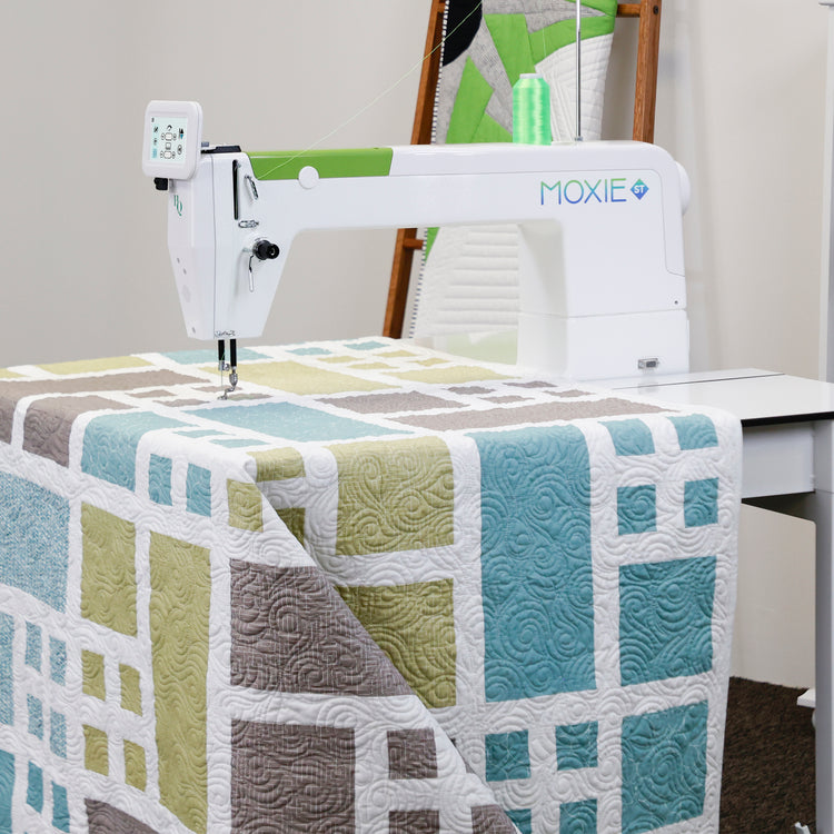 Moxie ST sewing machine is shown quilting a large patchwork quilt with geometric blocks in blue, sage green, taupe, and white. The sewing machine features a digital controller on the arm and the Moxie brand logo.