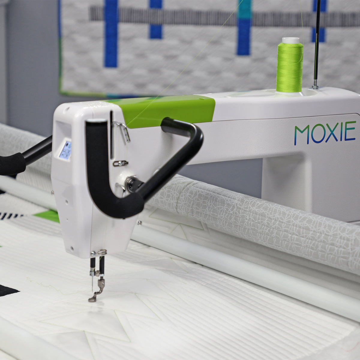 A close-up of a white Moxie quilting machine with a bright green spool of thread, stitching white fabric. The machine features a white body with a lime green accent strip and the brand name 'MOXIE' printed in blue and green on the side.