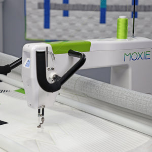 A close-up of a white Moxie quilting machine with a bright green spool of thread, stitching white fabric. The machine features a white body with a lime green accent strip and the brand name 'MOXIE' printed in blue and green on the side.