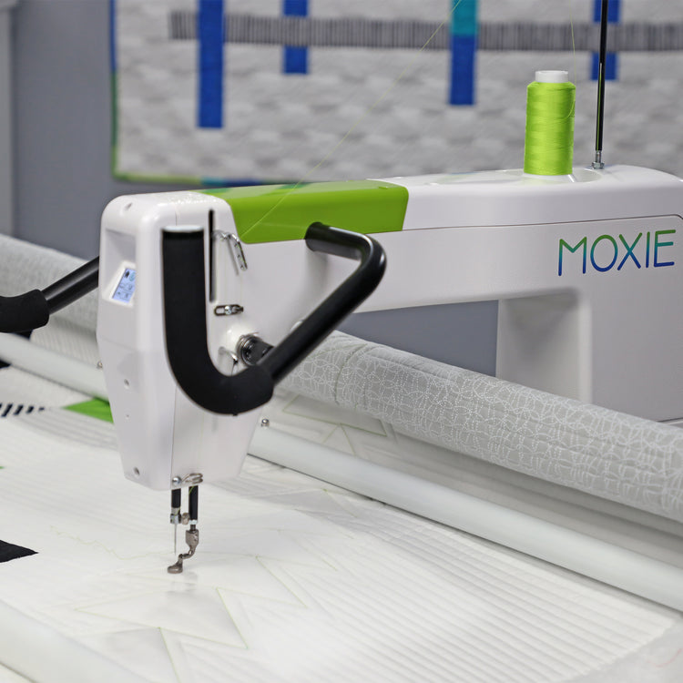 A close-up of a white Moxie quilting machine with a bright green spool of thread, stitching white fabric. The machine features a white body with a lime green accent strip and the brand name 'MOXIE' printed in blue and green on the side.