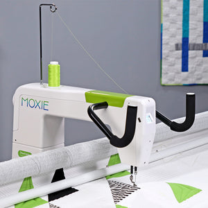 A white and lime green Moxie quilting machine with black handles is shown quilting a white fabric with green and black geometric patterns. A spool of lime green thread sits on top of the machine.