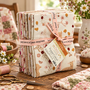 A stack of fabric tied with a pink ribbon, displayed on a rustic wooden table with sewing scissors, thread spools, and floral decor.