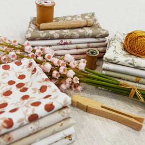 Folded fabric bundles in soft neutral and floral tones, arranged with wooden spools and pink blossoms on a light fabric surface.