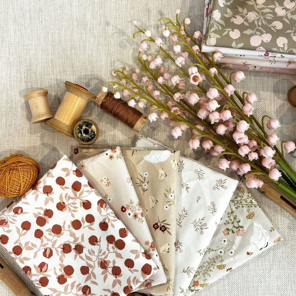 Fabric with botanical and fruit prints, arranged on a linen surface with spools of thread, a ball of yarn, and pink flowering stems.