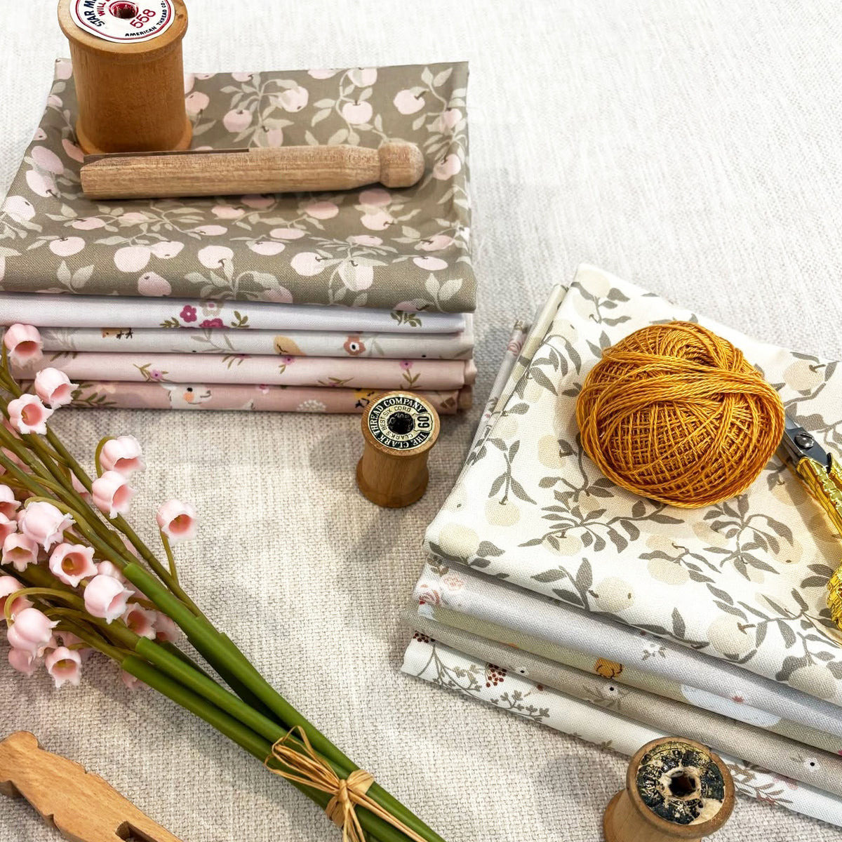 Folded patterned fabrics on a linen surface, with pink flowers in the foreground and a wooden spool handle resting on top.