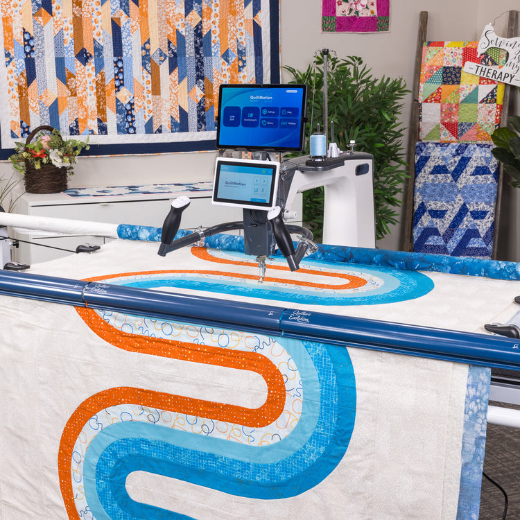 A large quilting machine with dual screens is shown stitching a quilt featuring a wavy blue and orange pattern on a white fabric. The setting appears to be a quilted craft studio with other quilts and plants in the background.