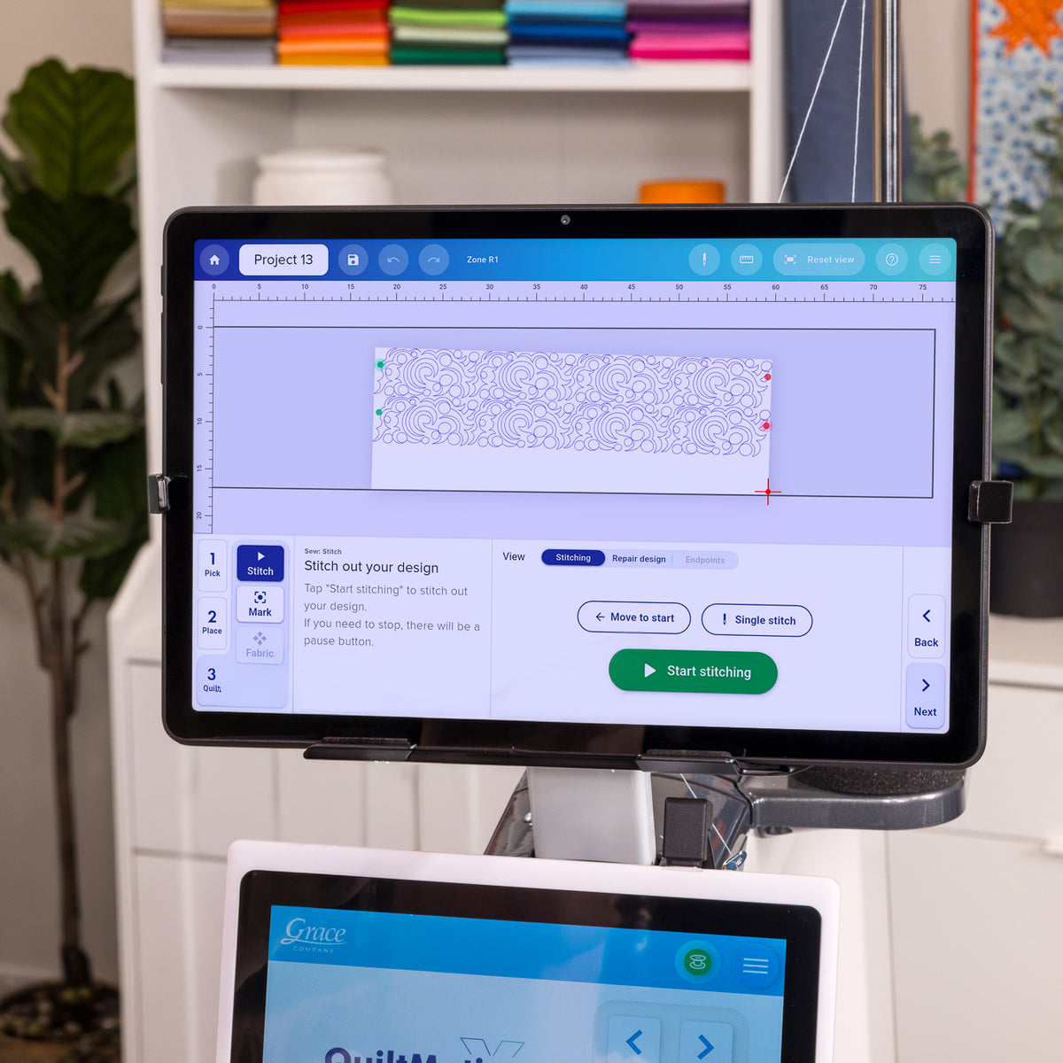 QMX Tablet with a large touchscreen display showing a design in progress, mounted on a stand in a bright indoor setting with shelves and plants in the background.