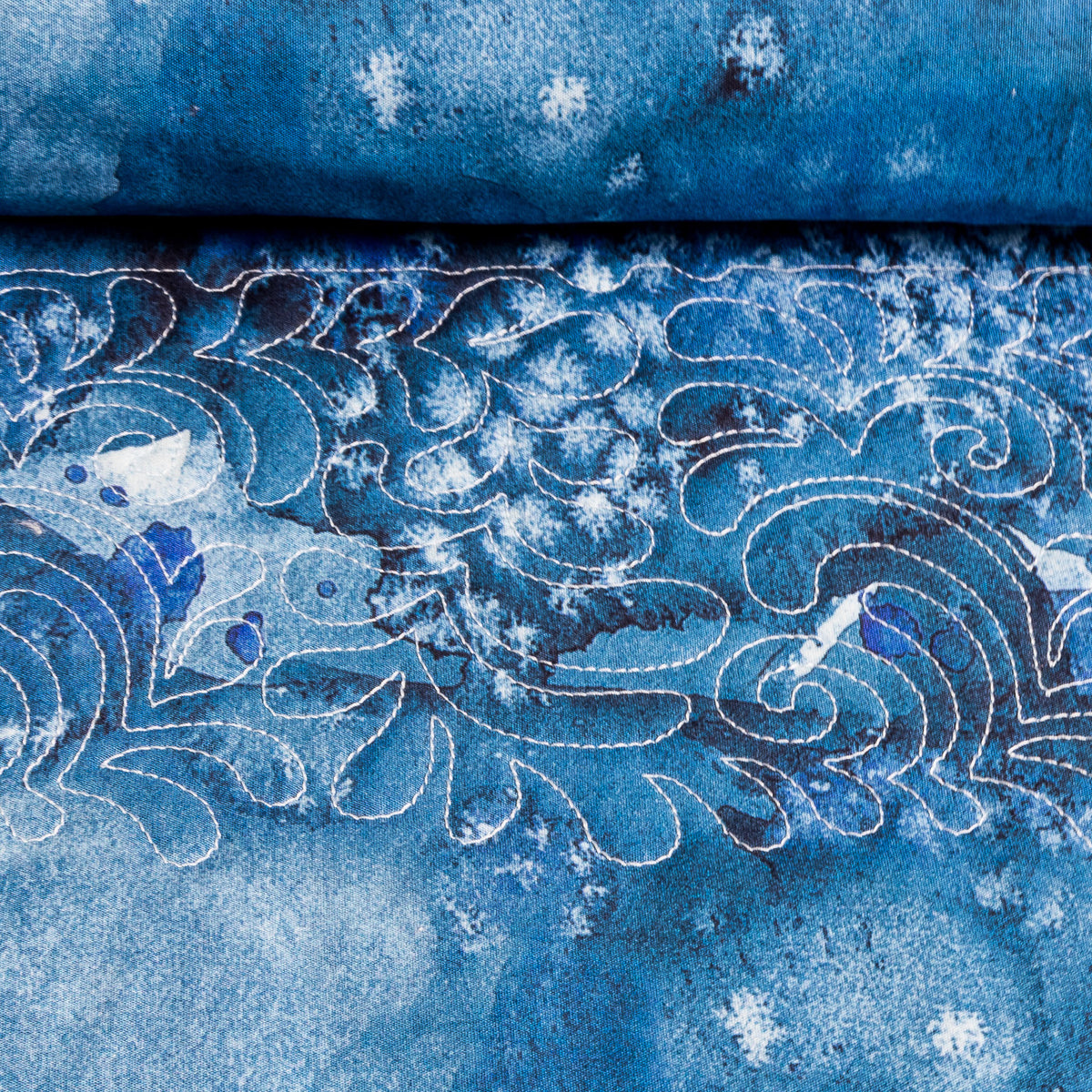 A close-up of blue patterned fabric with white stitched wave-like outlines and scattered white floral or snowflake-like motifs, with a folded section of the same fabric at the top.