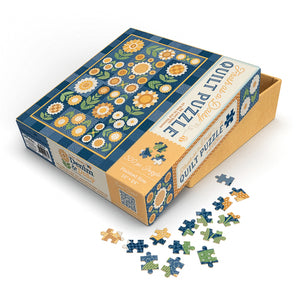 A jigsaw puzzle box with a floral quilt pattern, shown open with several loose puzzle pieces scattered in front on a clean white background.