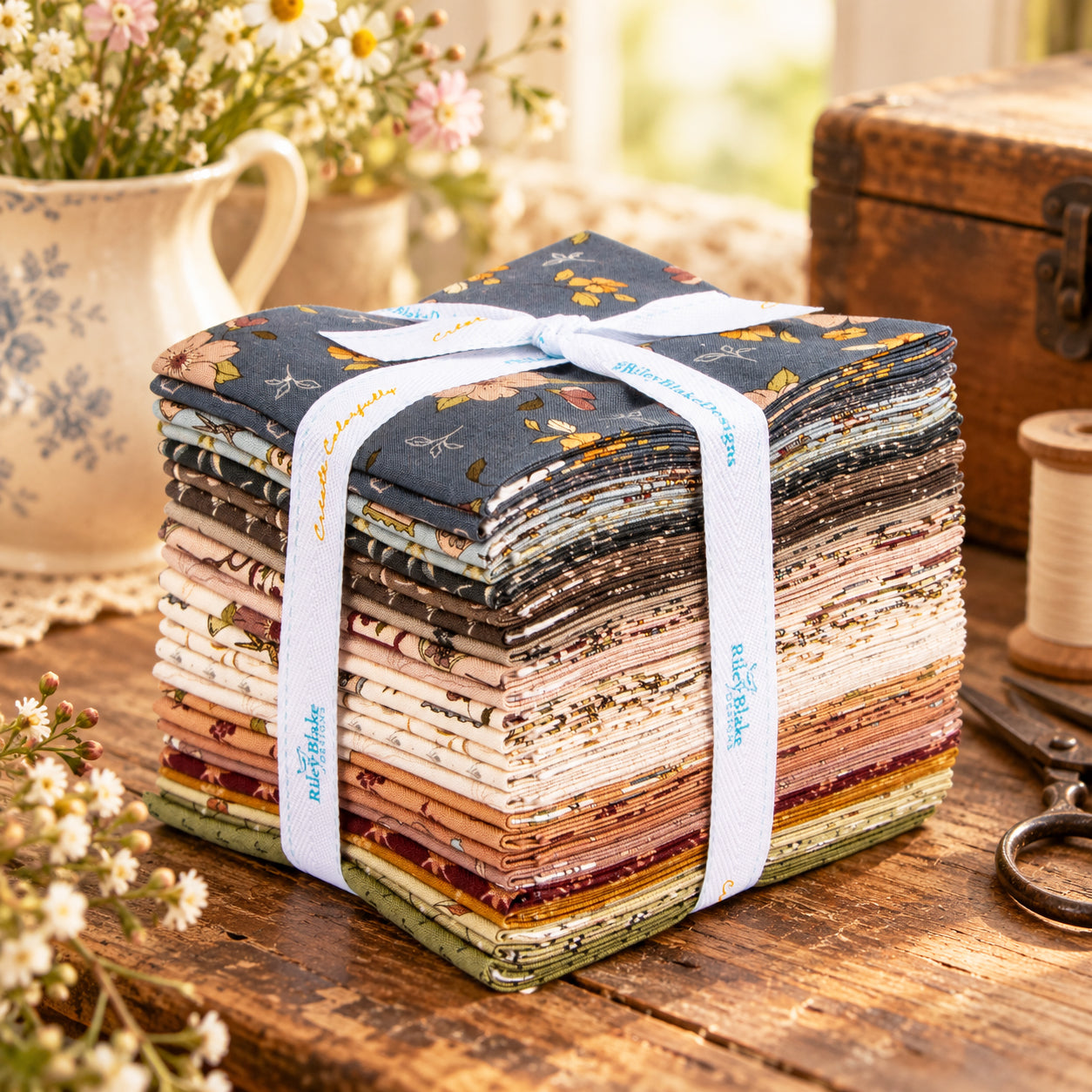 A stack of fabric cut pieces tied with a white ribbon, displayed on a rustic wooden table with flowers, scissors, and a spool in the background.