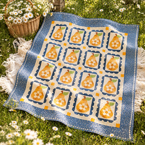 A quilt with a blue border and a cream center featuring yellow pears in floral frames, laid out on green grass with white daisies and a wicker basket nearby.