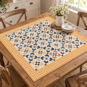 A square quilted table runner with a geometric patchwork pattern in navy blue, mustard yellow, and cream, displayed on a rustic wooden dining table with a vase of daisies and plates.