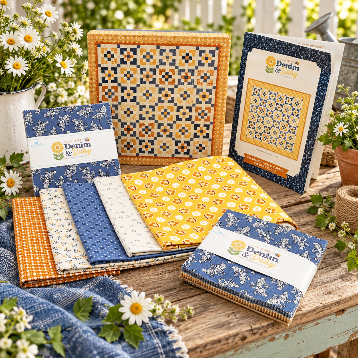 A quilt kit is displayed on a table with flowers and greenery around it. Visible items include folded fabric, a pattern and keepsake box.
