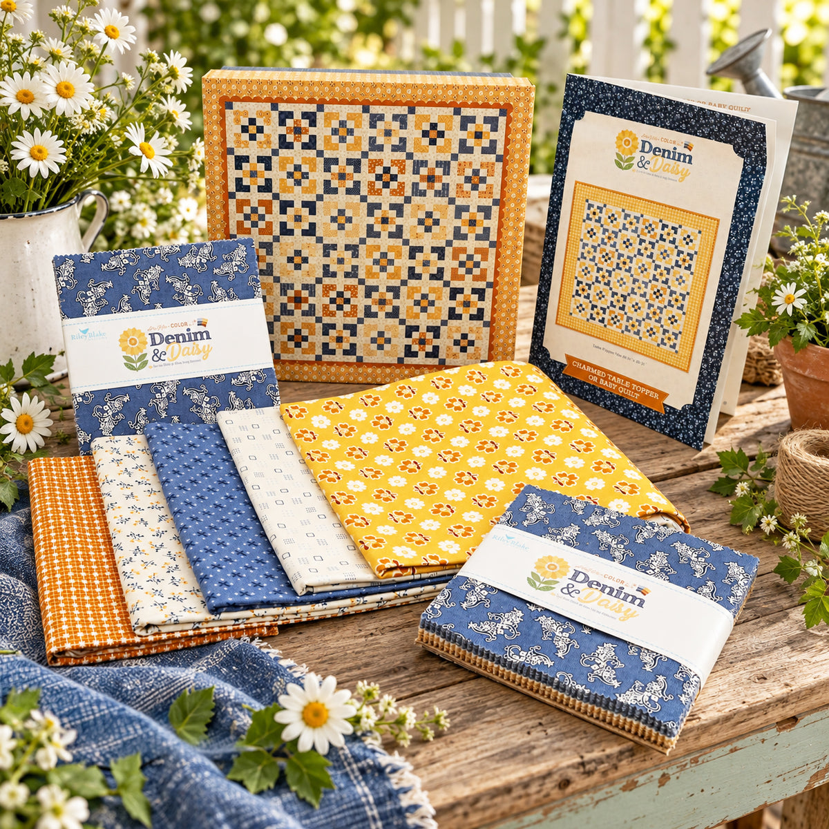 A quilt kit is displayed on a table with flowers and greenery around it. Visible items include folded fabric, a pattern and keepsake box.