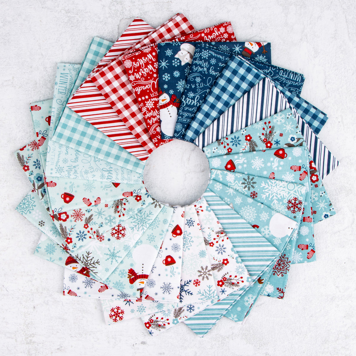 A Perfect Winter fabric assortment featuring festive snowmen, snowflakes, mittens, winter florals, holiday lettering, gingham checks, and stripes in icy blue, navy, red, and white, arranged in a circular fan.