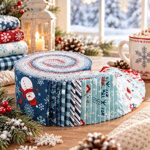 A Perfect Winter Rolie Polie fabric roll featuring snowmen, snowflakes, winter florals, gingham checks, and stripes in navy, icy blue, red, and white, displayed on a cozy holiday table with pinecones, evergreen branches, a glowing lantern, and a festive mug.