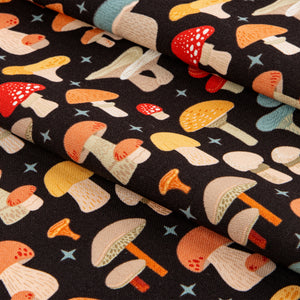 A close-up of a folded black fabric with a colorful mushroom print, showing various mushroom illustrations in orange, yellow, red, and beige, accented with small blue stars.