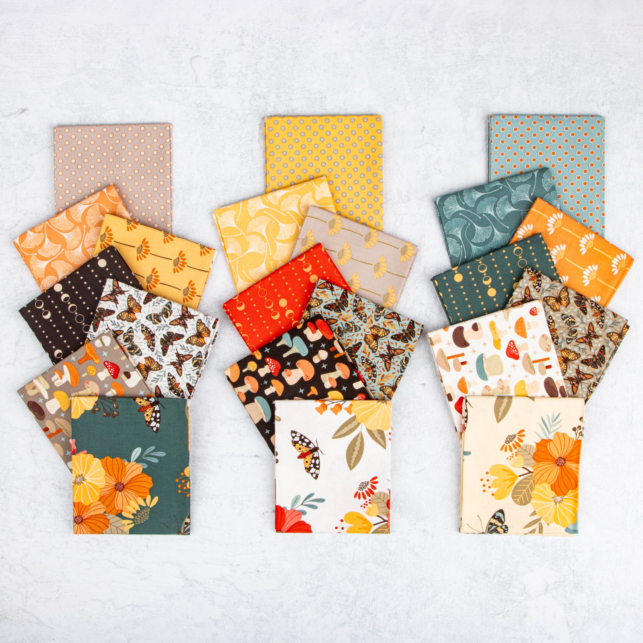 Three stacks of patterned fabric squares arranged side by side on a light textured background. The fabrics feature autumn-themed prints including orange pumpkins, butterflies, leaves, mushrooms, and floral motifs in warm tones like orange, yellow, teal, brown, cream, and red.