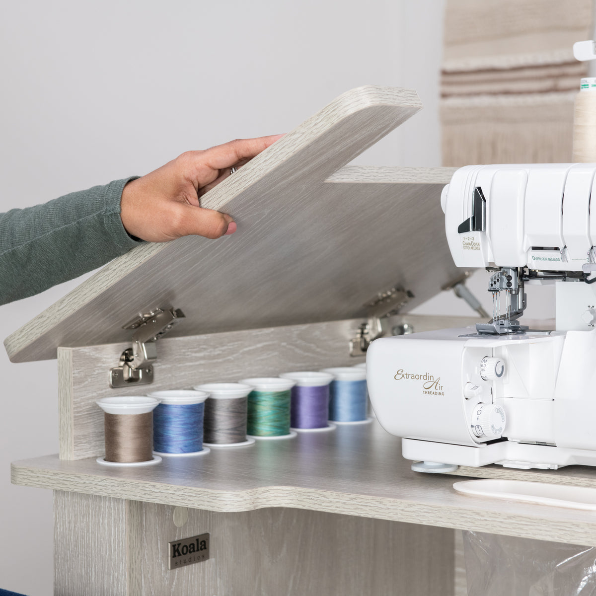 Person lifting the hinged lid of a Koala Serger Studio cabinet, revealing thread spool storage and serger