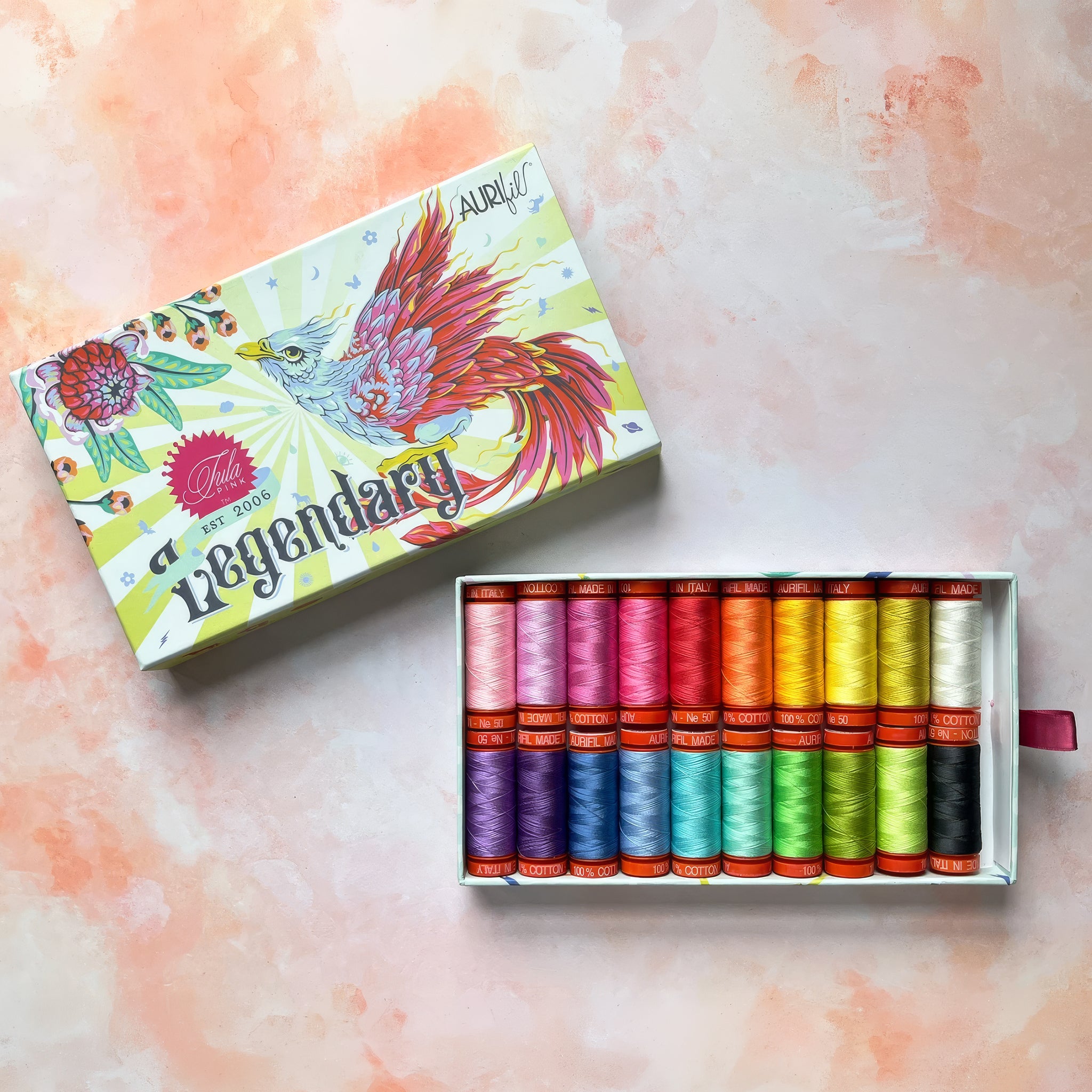 A boxed set of multicolored sewing thread spools displayed next to its illustrated lid on a soft peach-pink textured background. The box is labeled 'Legendary' and 'Aurifil'.