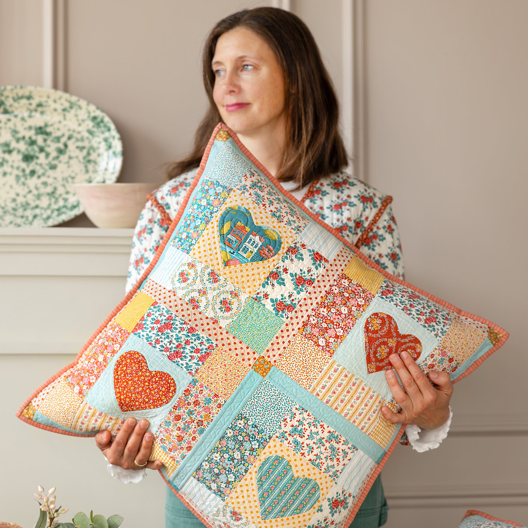 Woman holding a patchwork Village Life pillow with heart appliqués and colorful florals.