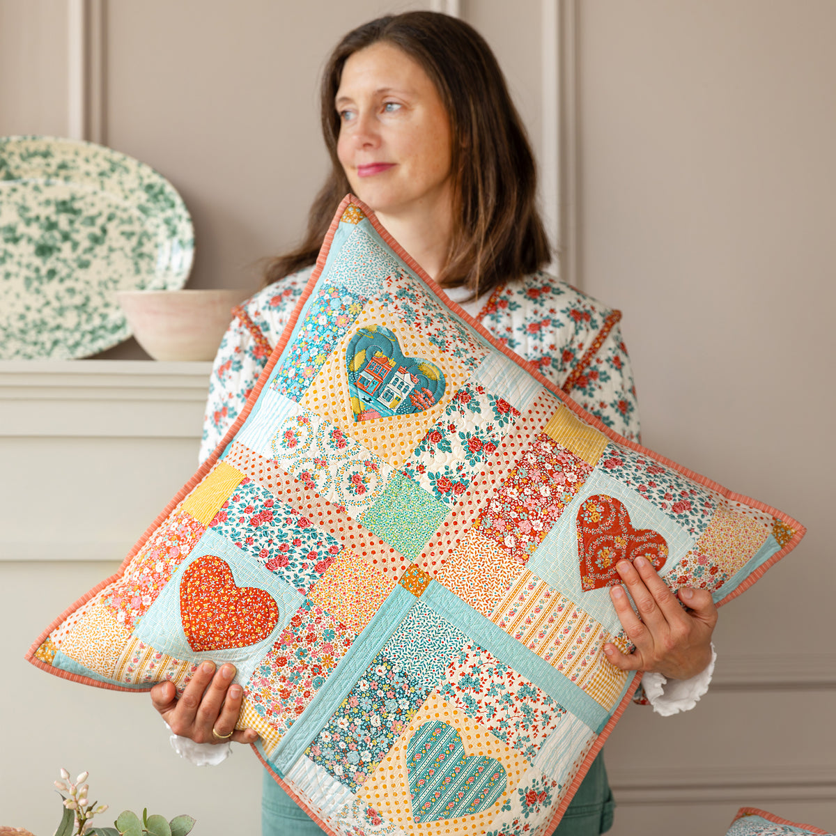Woman holding a patchwork Village Life pillow with heart appliqués and colorful florals.
