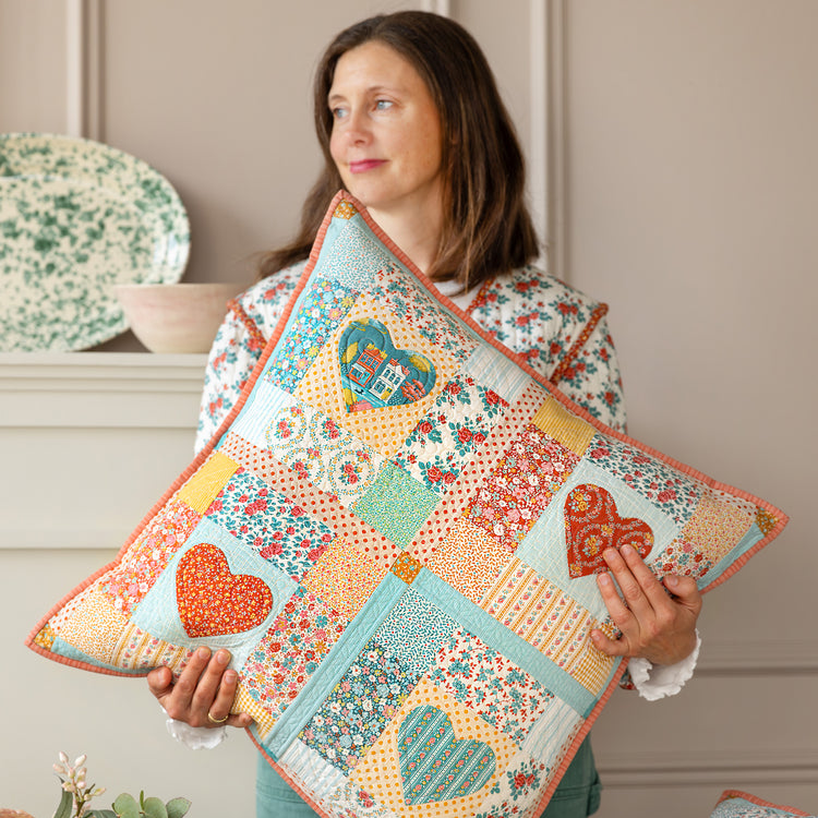 Woman holding a patchwork Village Life pillow with heart appliqués and colorful florals.